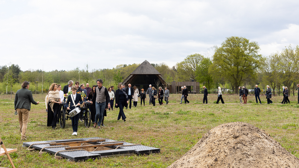 Rouwstoet met trommelaar voorop, over een veld op de natuurbegraafplaats, met het open graf in de voorgrond